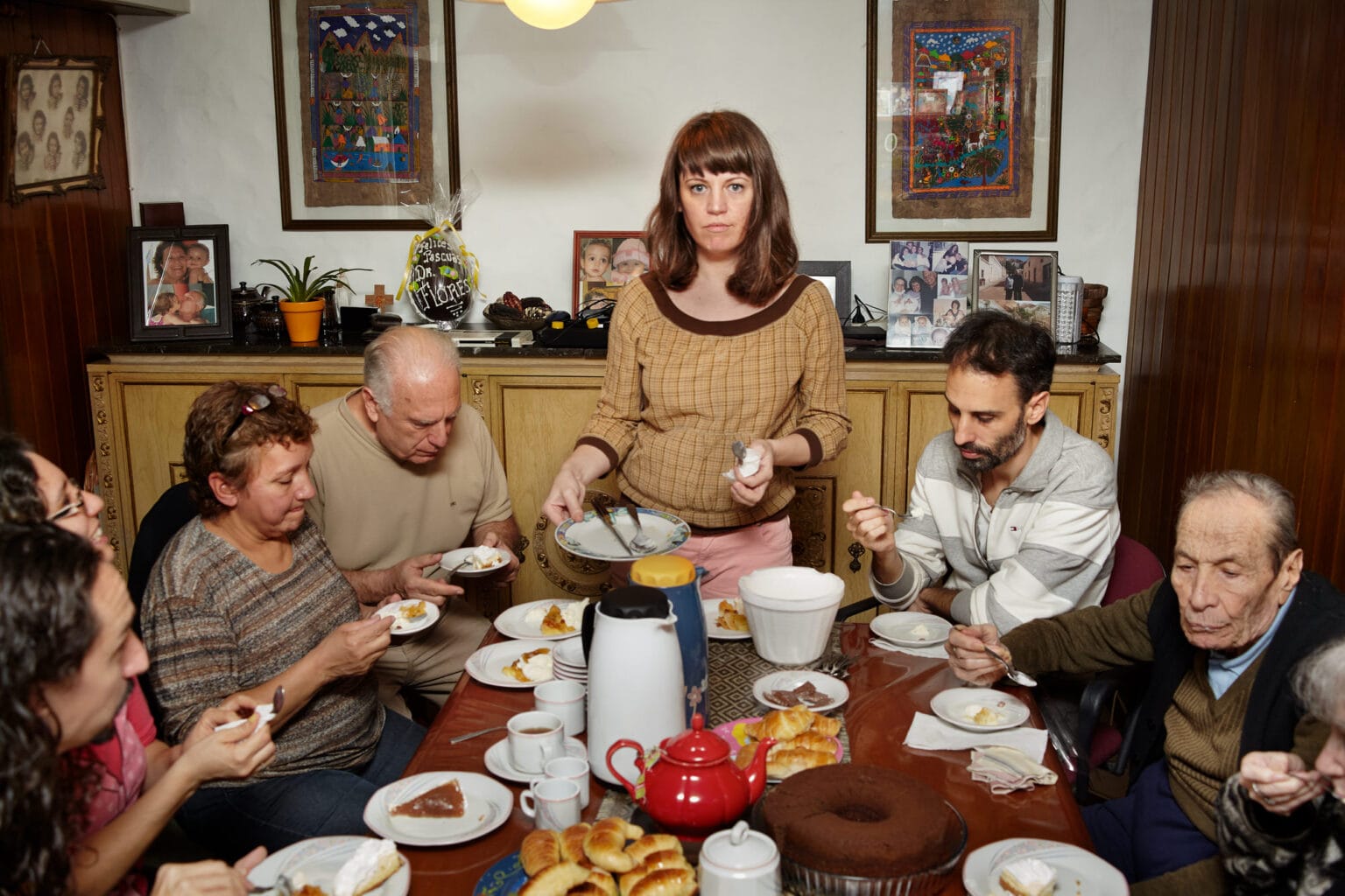 Woman named Gaviota serves dessert at a crowded Flowers family gathering table, surrounded by relatives enjoying coffee, cake, and pastries in a cozy dining room decorated with framed photos and colorful artwork
