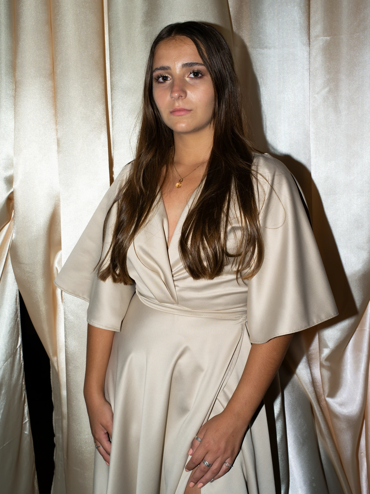 Young woman standing in front of beige satin curtains, wearing a light champagne wrap dress, photographed with direct flash in a studio-style portrait. The Graduates series, Cecilia Reynoso.