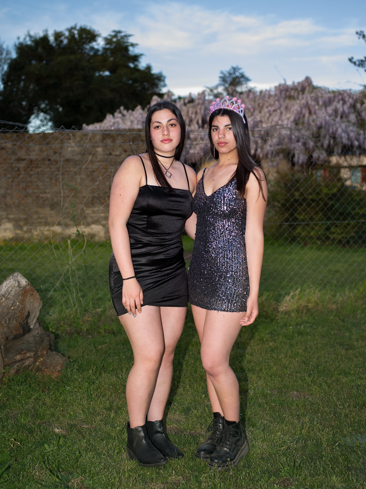 Two young women posing outdoors on grass in short party dresses, one wearing a tiara, photographed with flash against a chain-link fence and trees. The Graduates series, Cecilia Reynoso.