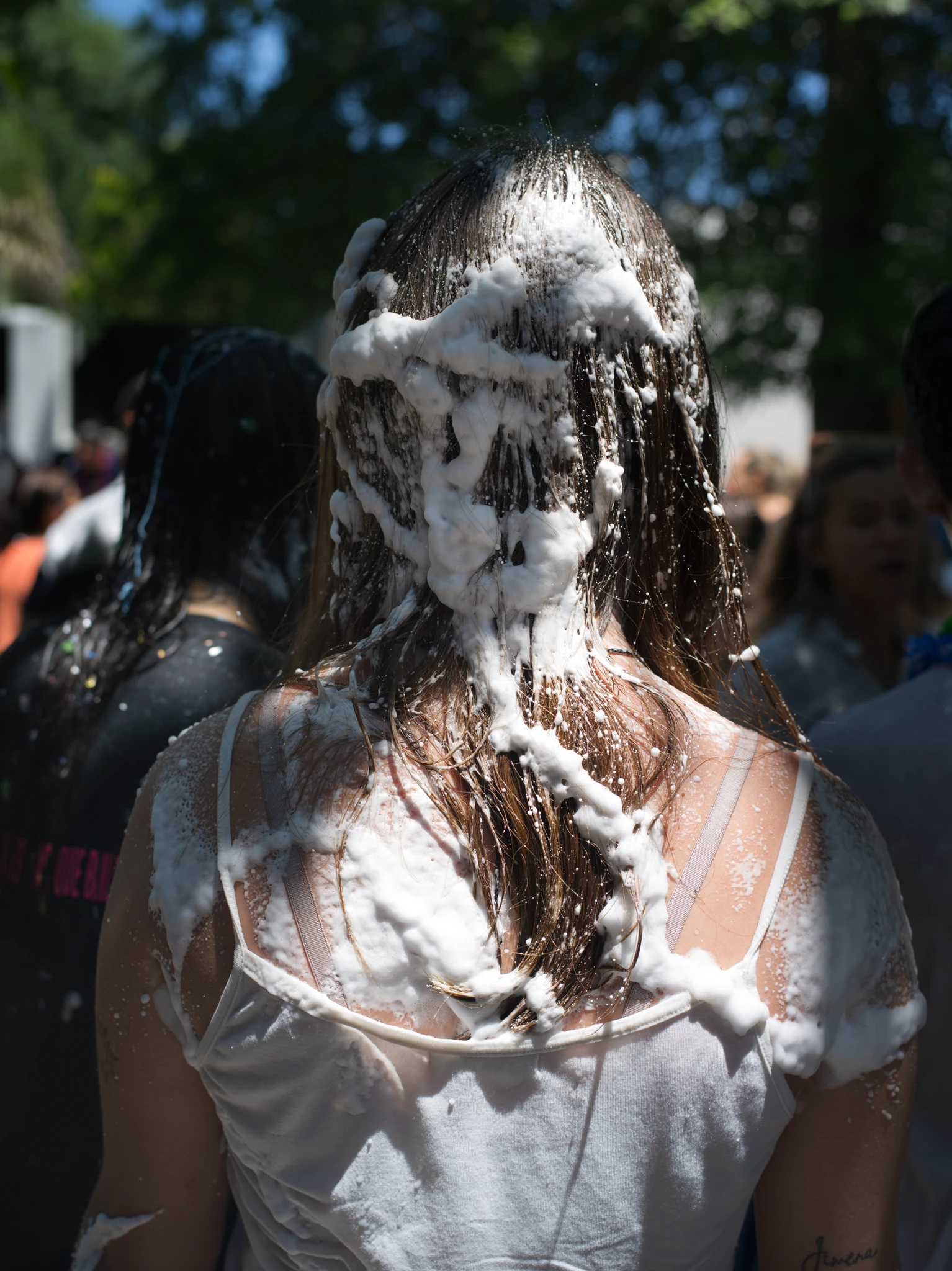 Back view of a young woman in a crowd with white foam covering her hair and shoulders during a public celebration, photographed outdoors in daylight. The Graduates series, Cecilia Reynoso.