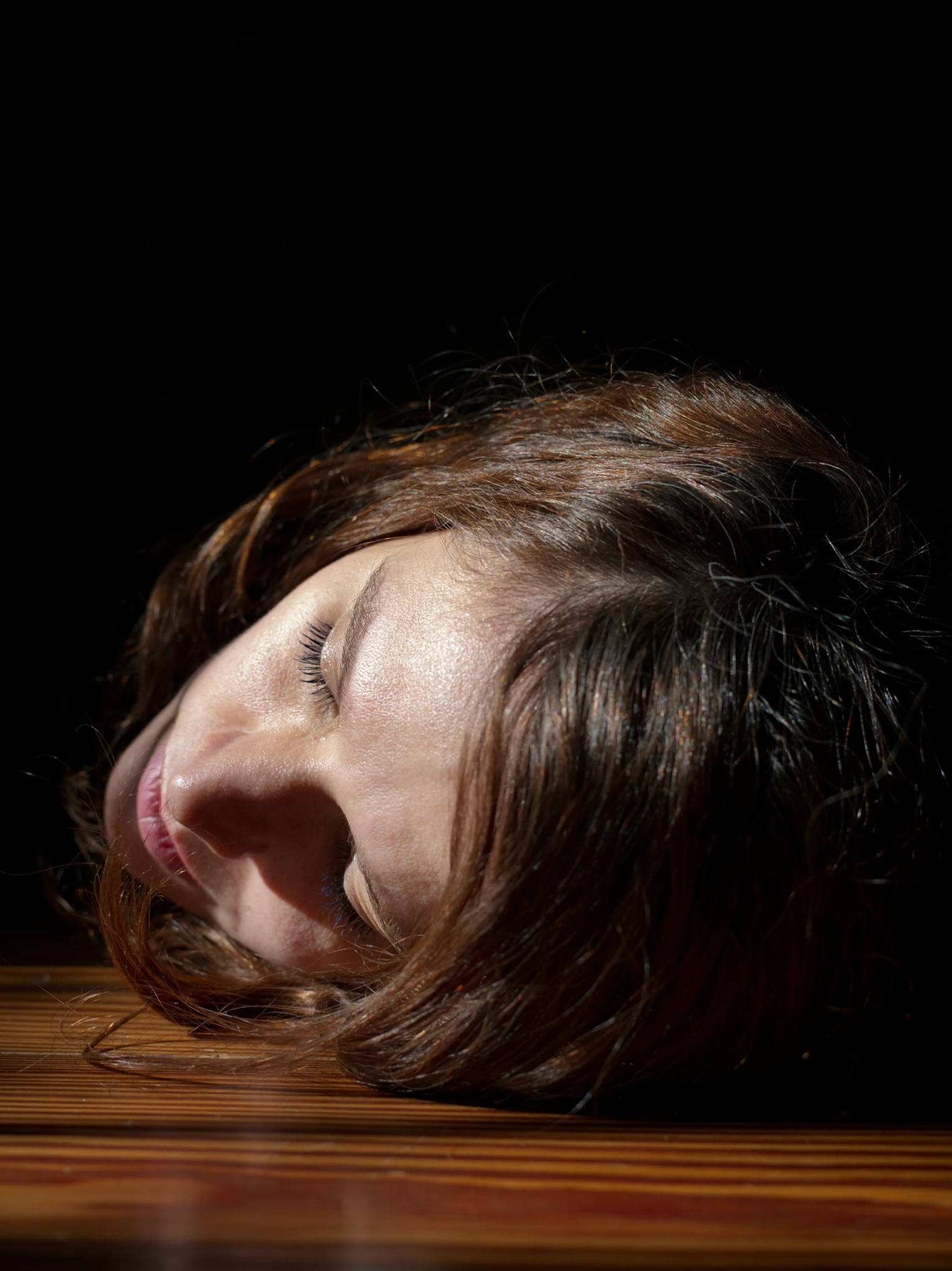 Close-up of a woman lying on her left side  with eyes closed, her face resting on a wooden floor against a black background, To forget 2020, a short log of the pandemic in Buenos Aires series, Buenos Aires, 2020.